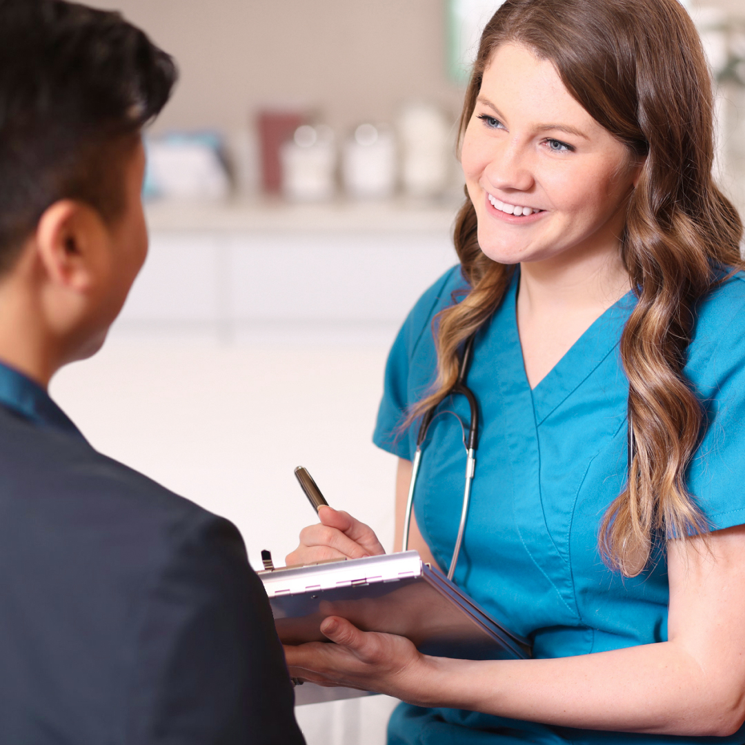 An RN holding a clipboard and pen talking to a patient in an office.