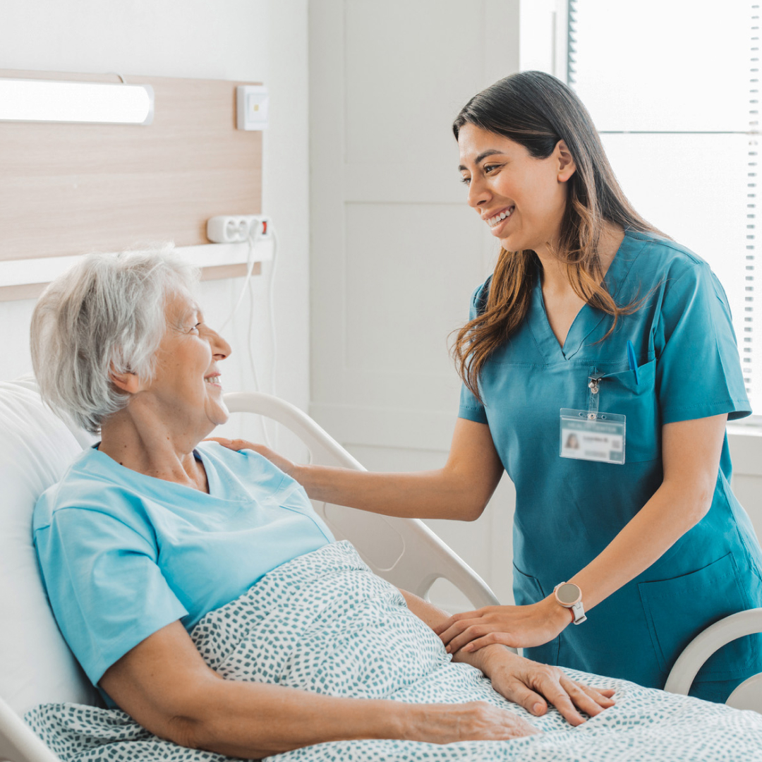 A nurse smiling at and putting her hand on the shoulder of a client sitting upright in a hospital bed.