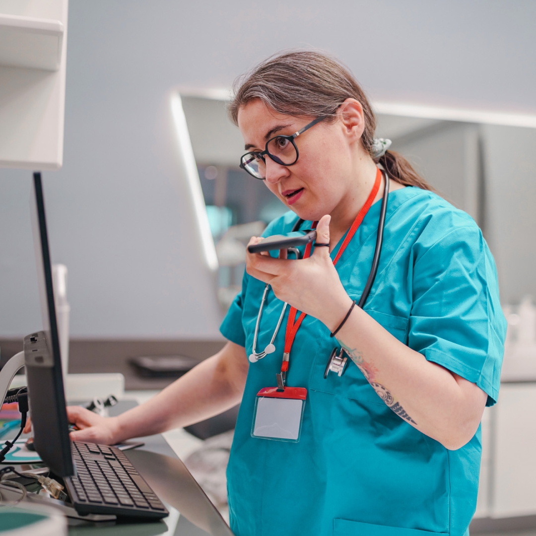 A nurse using a computer and speaking into a smartphone.