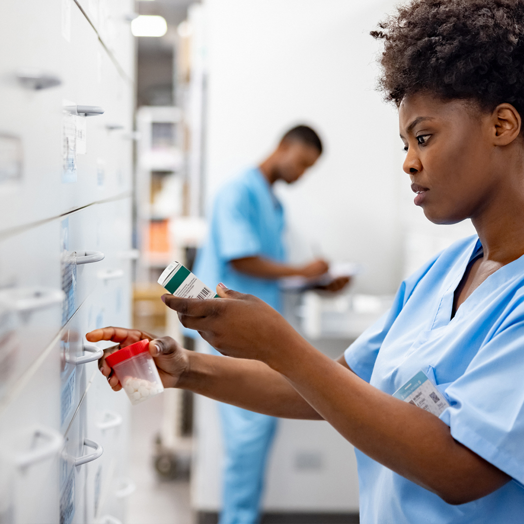 Nurse wearing scrubs selecting medication from a storage cabinet while reviewing a prescription label in a clinical setting.