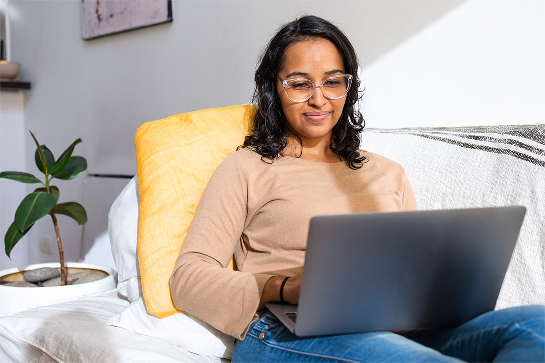 A South Asian nurse wearing casual clothes using a laptop at home with a smile on her face.