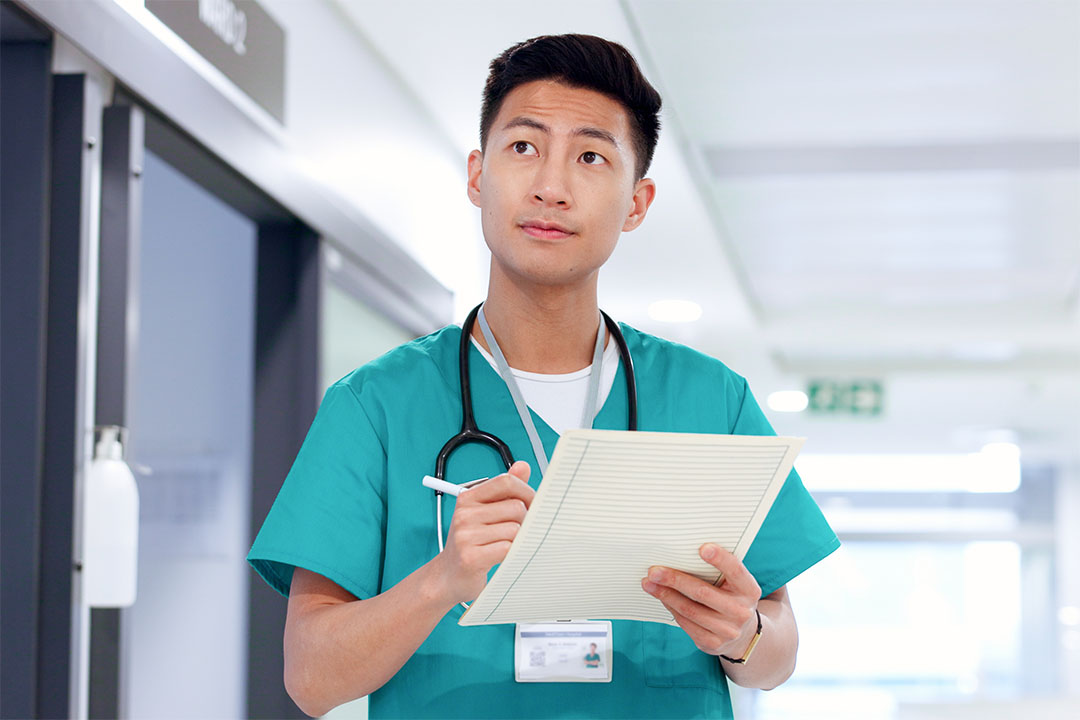 An East Asian male nurse wearing scrubs in a hospital corridor holding paperwork and looking up in deep thought.
