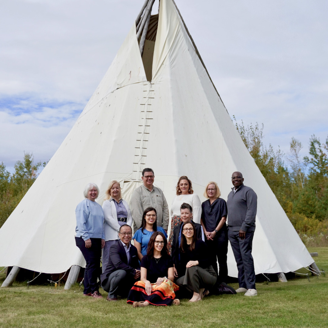 Dix personnes sont debout et agenouillées devant un grand tipi blanc dans un champ gazonné. Des arbres entourent l'endroit et un ciel nuageux est visible à l'arrière-plan.